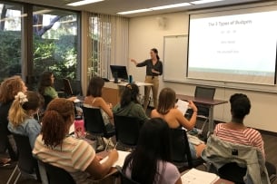 A Scripps College alumna teaches a small class, with a projector screen stating that the lesson is about the three types of budgeting.